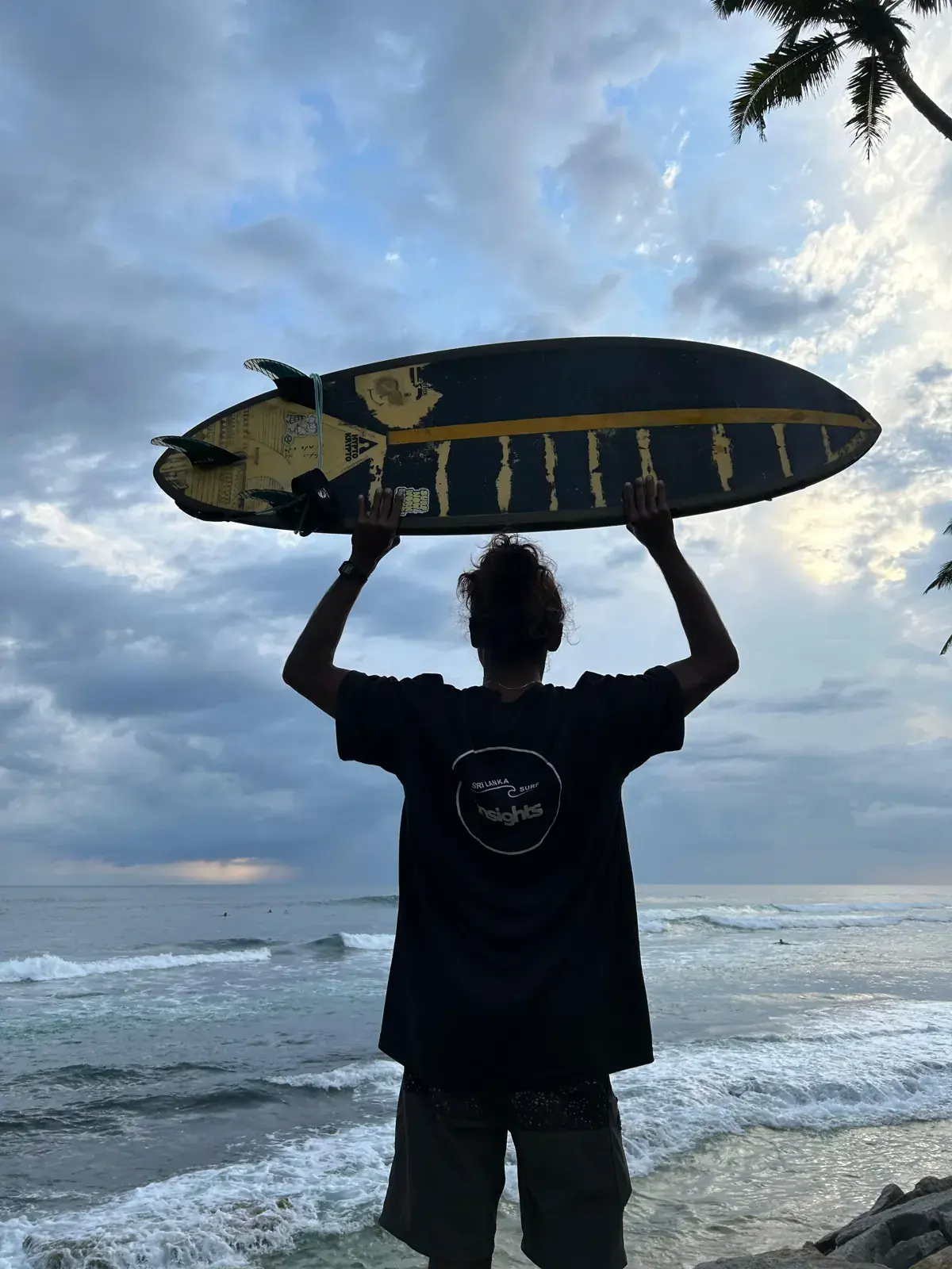 Surfer holding a surfboard overhead, facing the sea under a cloudy sky