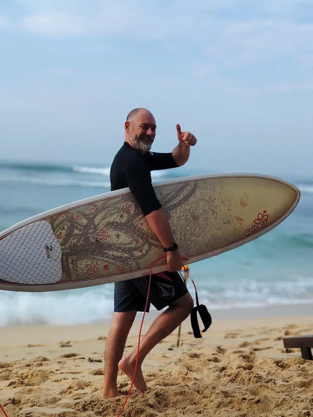 Smiling guest giving a thumbs-up with a surfboard on the beach