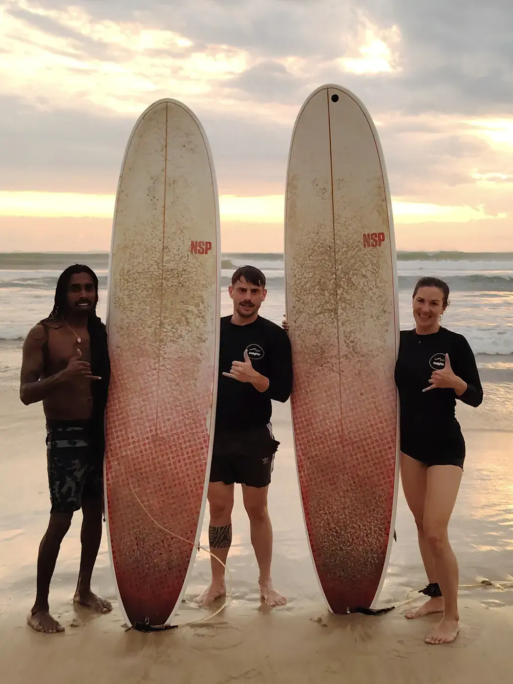 Three surfers standing with tall surfboards on the beach at sunset