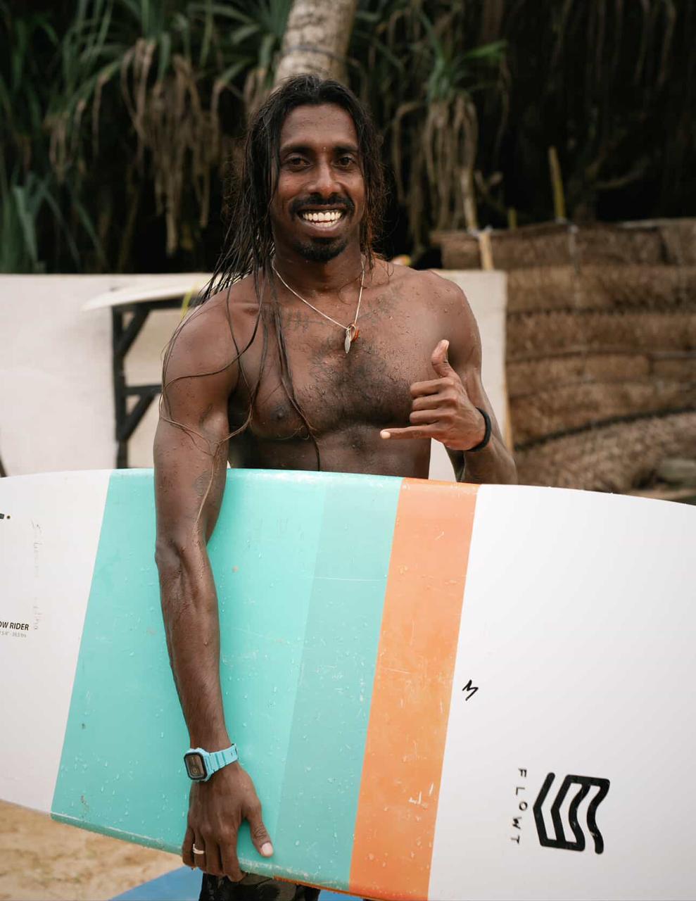 Ravi holding a surfboard and giving a shaka sign